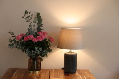 Photo of Beautiful bouquet of roses and eucalyptus branches in vase near lamp on wooden table indoors