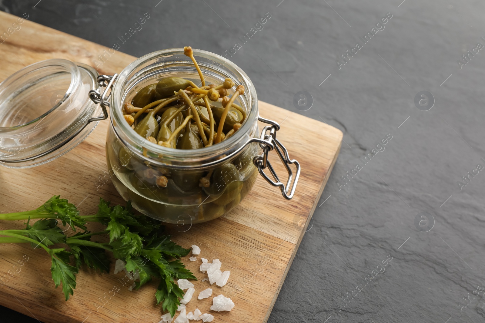 Photo of Tasty capers in glass jar, salt and parsley on black table, space for text