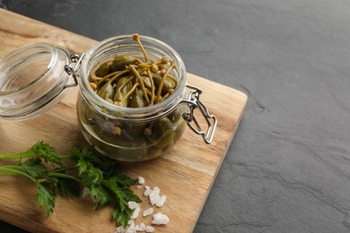 Photo of Tasty capers in glass jar, salt and parsley on black table, space for text