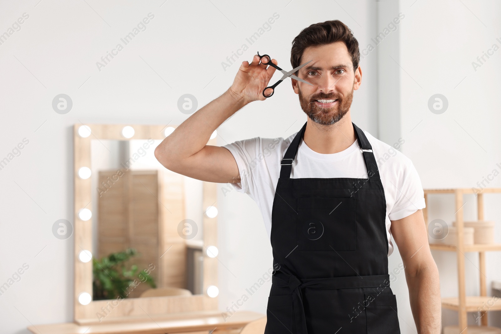 Photo of Smiling hairdresser in apron holding scissors near vanity mirror in salon, space for text