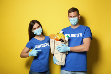 Photo of Volunteers in protective masks and gloves with products on yellow background. Aid during coronavirus quarantine