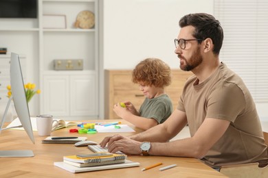 Photo of Man working remotely at home. Father using computer while his son playing at desk
