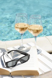 Photo of Glasses of tasty wine, sunglasses and open book on swimming pool edge