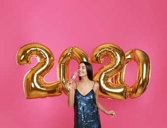 Photo of Happy young woman with glass of champagne near golden 2020 balloons on pink background. New Year celebration