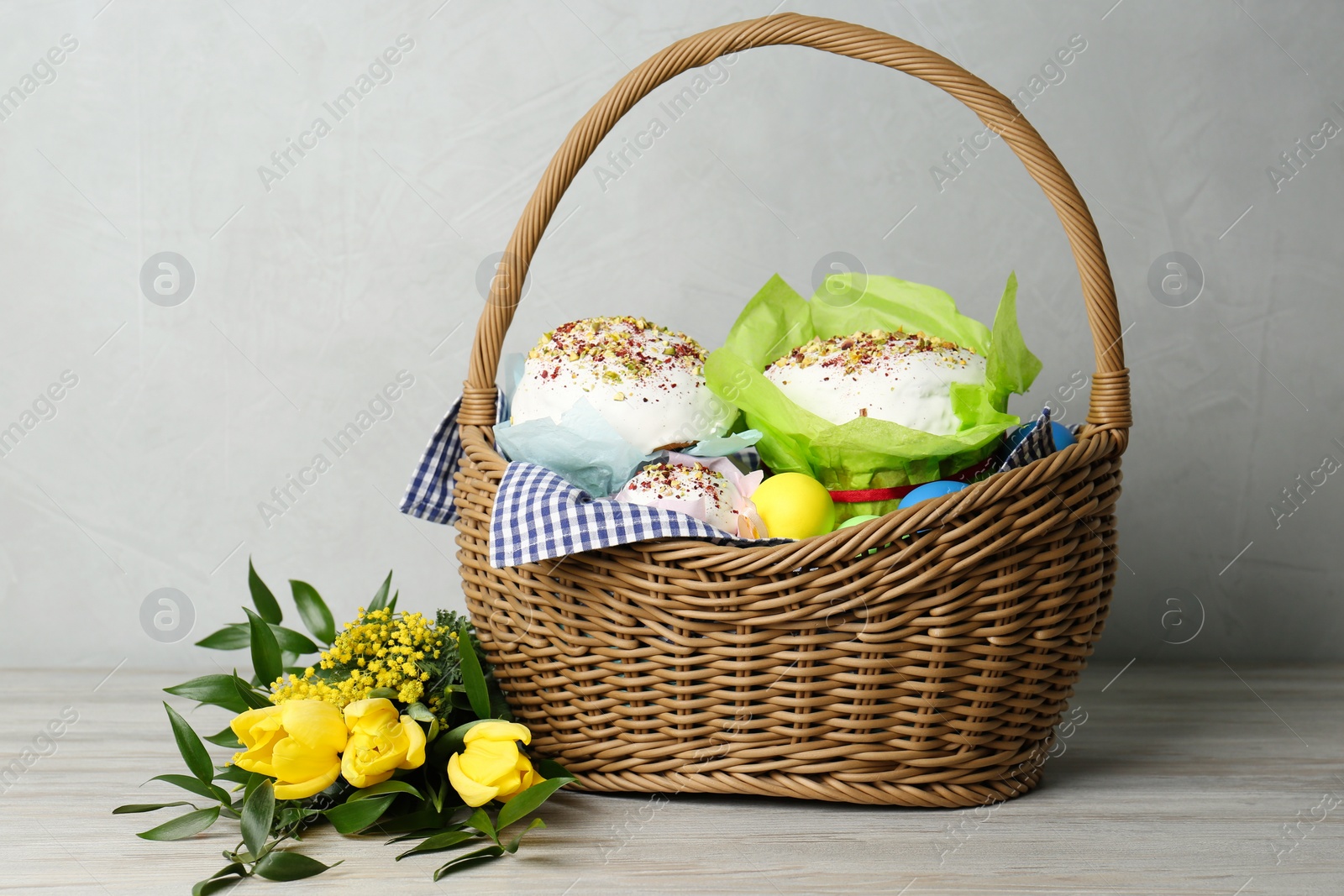 Photo of Basket with delicious Easter cakes, dyed eggs and flowers on light wooden table