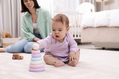 Photo of Cute baby girl playing with toy pyramid near mother on floor at home