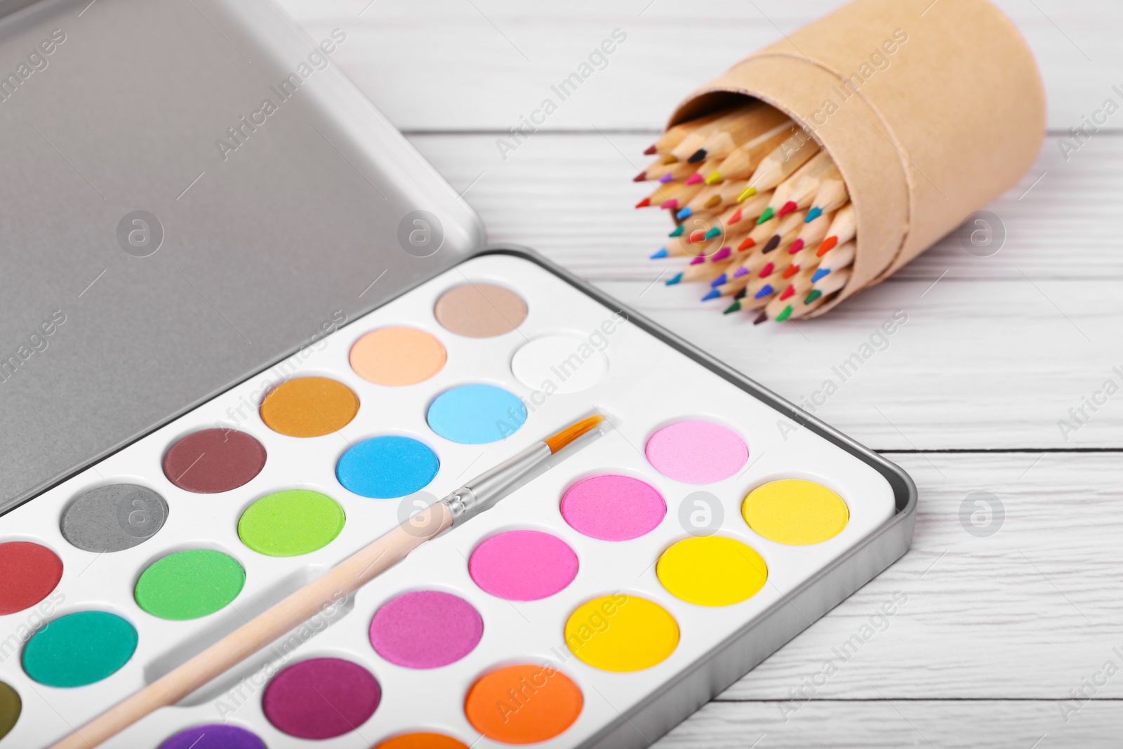 Photo of Watercolor palette with brush and colorful pencils on white wooden table, closeup