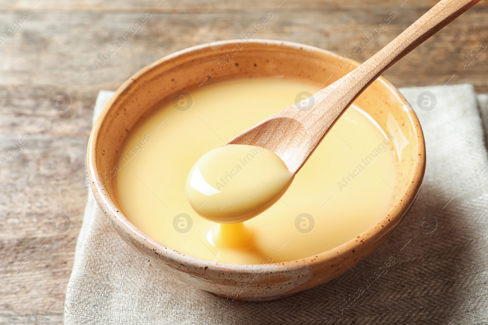 Photo of Spoon with pouring condensed milk over bowl on table, closeup. Dairy products