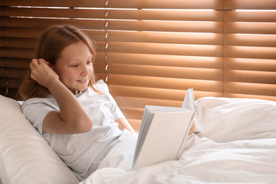 Photo of Cute preteen girl reading book in bed near window