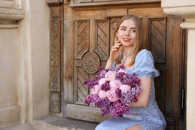 Beautiful woman with bouquet of spring flowers near building outdoors, space for text