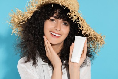 Photo of Beautiful happy woman in straw hat holding tube of sun protection cream on light blue background