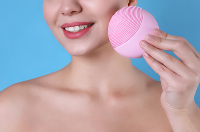 Photo of Young woman washing face with cleansing brush on light blue background, closeup. Cosmetic product