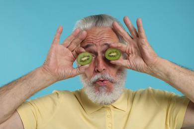 Photo of Senior man covering eyes with halves of kiwi on light blue background