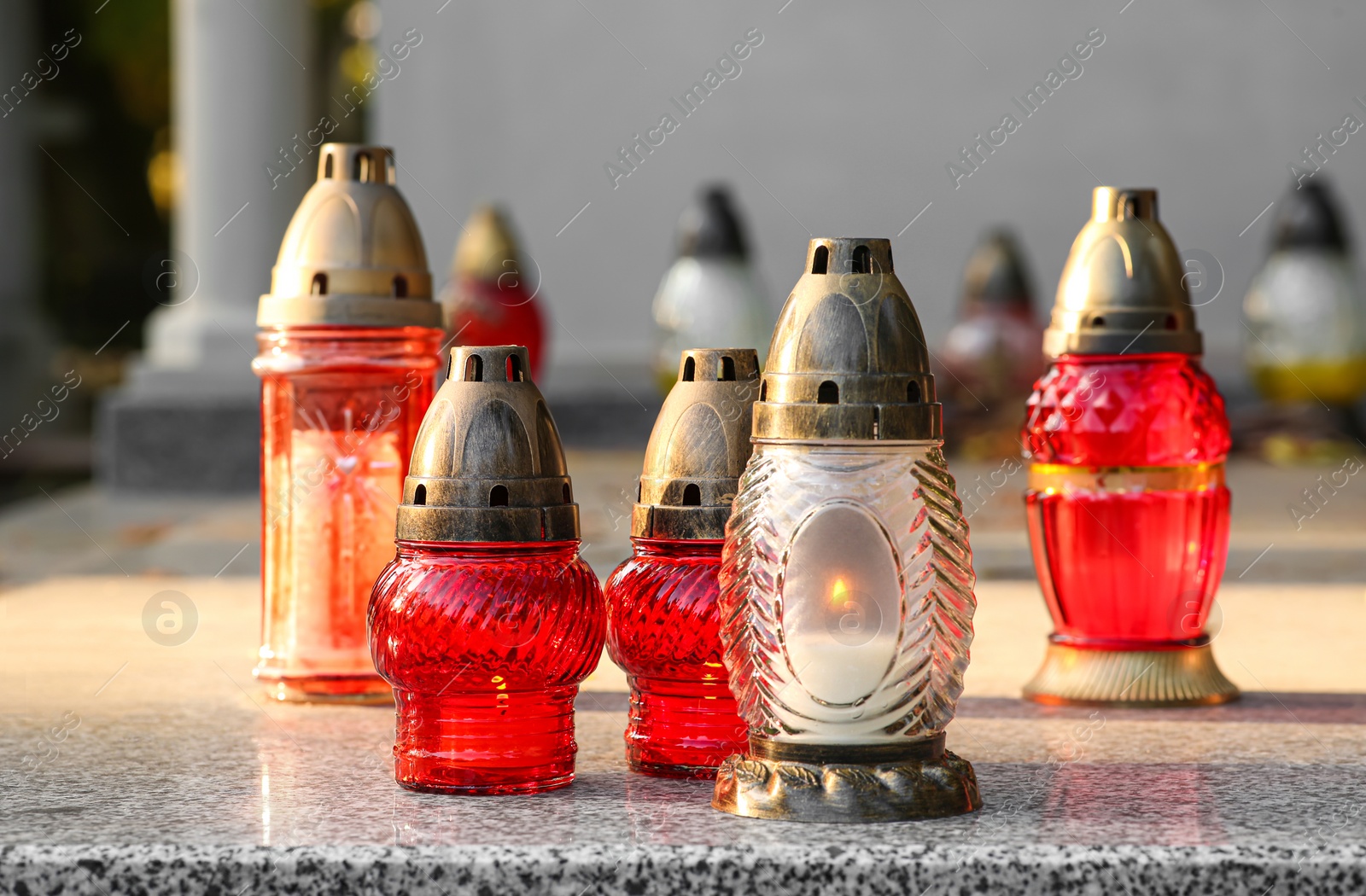 Photo of Different grave lanterns with burning candles on granite tombstone outdoors. Funeral ceremony