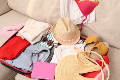 Open suitcase with different women clothes and accessories on sofa