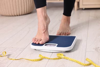 Photo of Woman stepping on floor scale and measuring tape at home, closeup. Weight control
