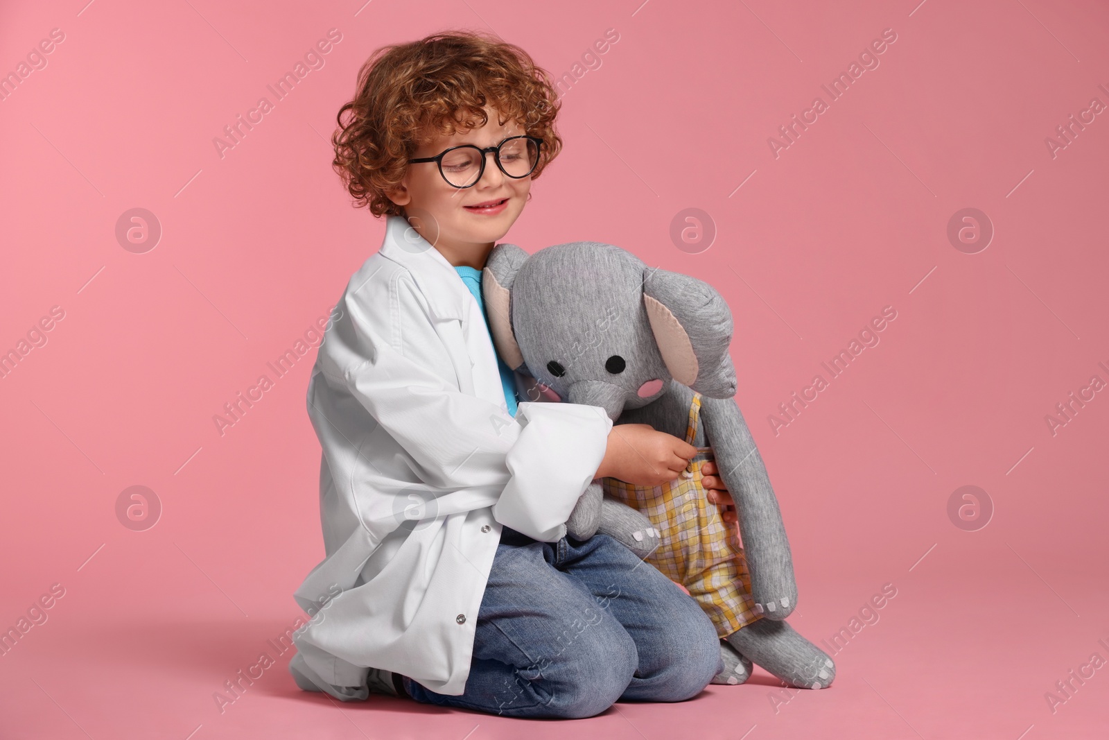 Photo of Little boy in medical uniform with toy elephant on pink background