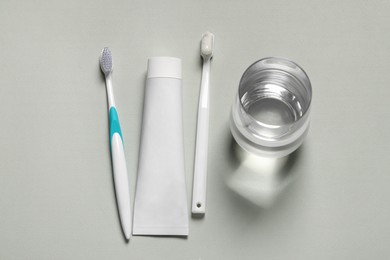 Photo of Plastic toothbrushes with paste and glass of water on grey background, flat lay