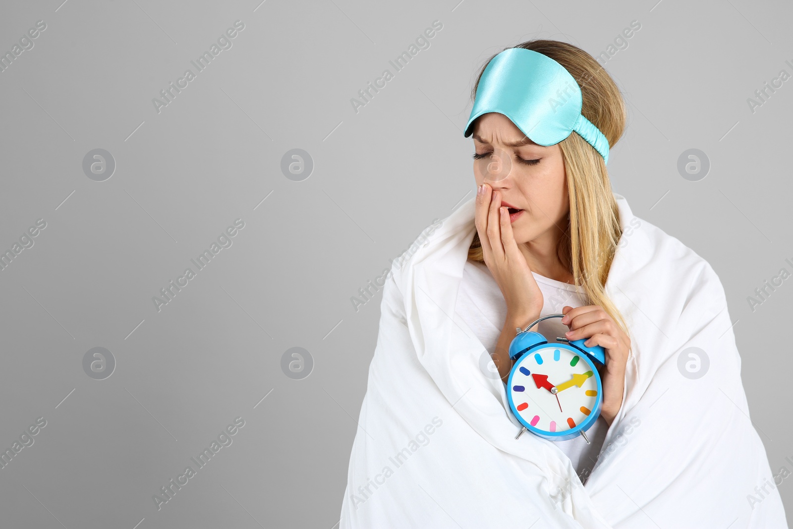 Photo of Woman in sleeping mask wrapped with blanket holding alarm clock on grey background. Space for text