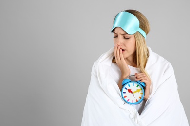 Woman in sleeping mask wrapped with blanket holding alarm clock on grey background. Space for text