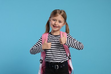 Happy schoolgirl with backpack showing thumbs up gesture on light blue background