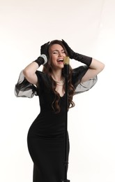 Beautiful young woman in stylish black dress with microphone singing on light background