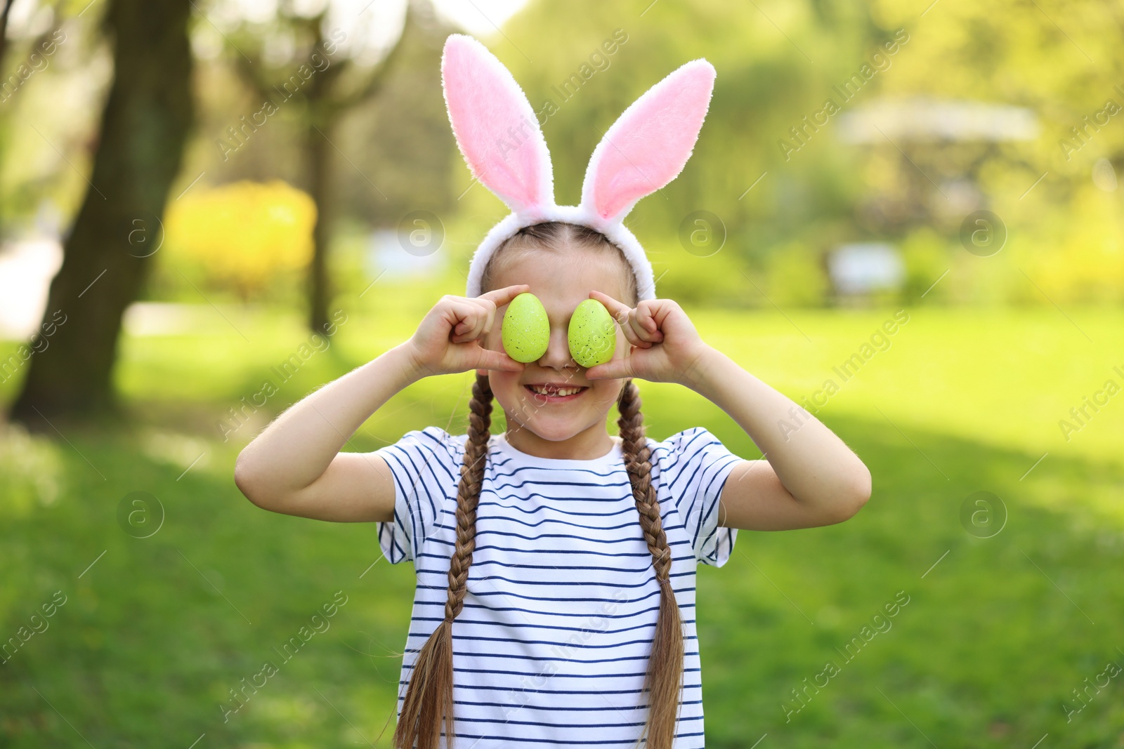 Photo of Easter celebration. Cute little girl in bunny ears covering eyes with painted eggs outdoors