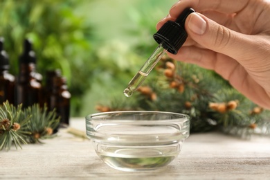 Photo of Woman holding pipette with conifer essential oil over bowl on wooden table, closeup