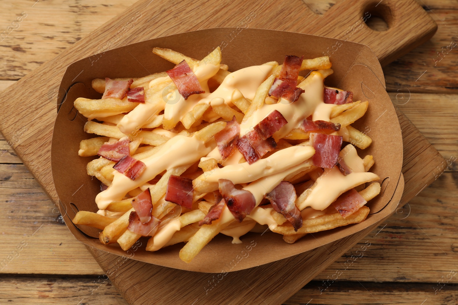 Photo of Tasty potato fries, cheese sauce and bacon in paper container on wooden table, top view