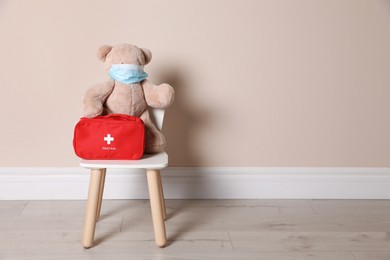 Photo of Toy bear with face mask and first aid bag near beige wall, space for text. Pediatrician practice