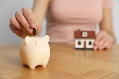 Photo of Woman putting money into piggy bank and holding little house model at wooden table, focus on hand