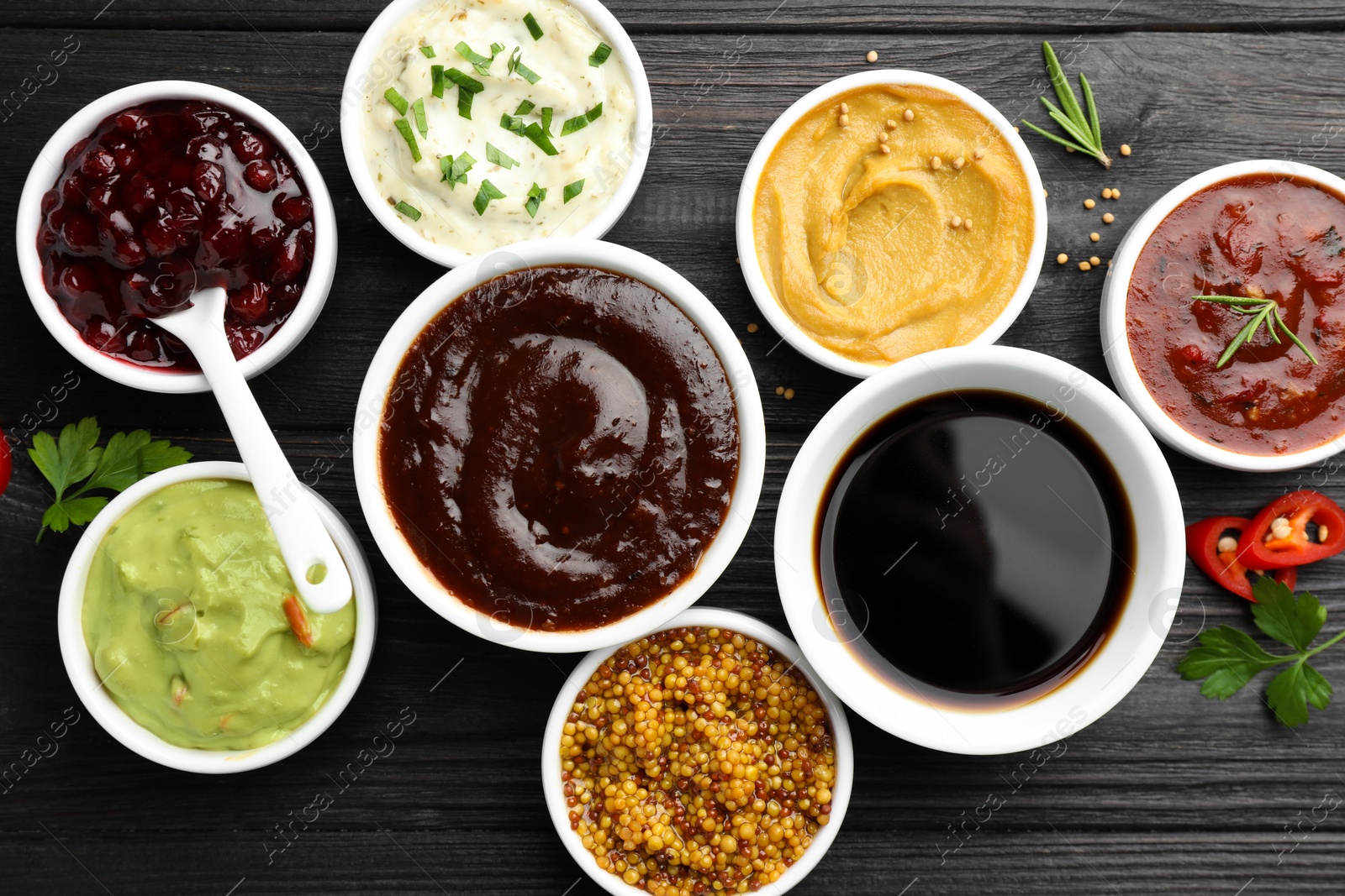 Photo of Different tasty sauces in bowls, parsley, chili pepper and rosemary on black wooden table, flat lay