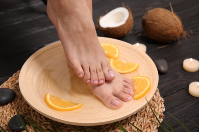 Photo of Woman soaking her feet in plate with water and orange slices on wooden floor, closeup. Spa treatment