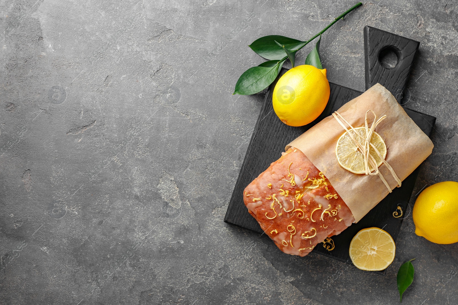 Photo of Wrapped tasty lemon cake with glaze and citrus fruits on grey textured table, flat lay. Space for text