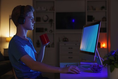 Photo of Teenage boy with cup of drink using computer in room at night. Internet addiction