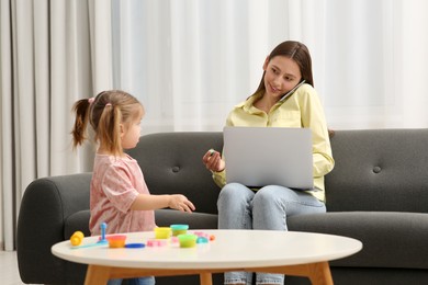 Photo of Little girl bothering her mother at home. Woman working remotely in living room