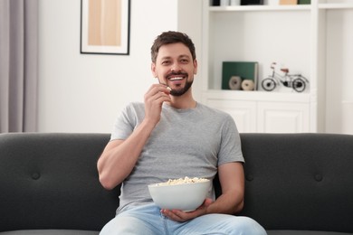 Happy man with bowl of popcorn watching movie via TV on sofa at home