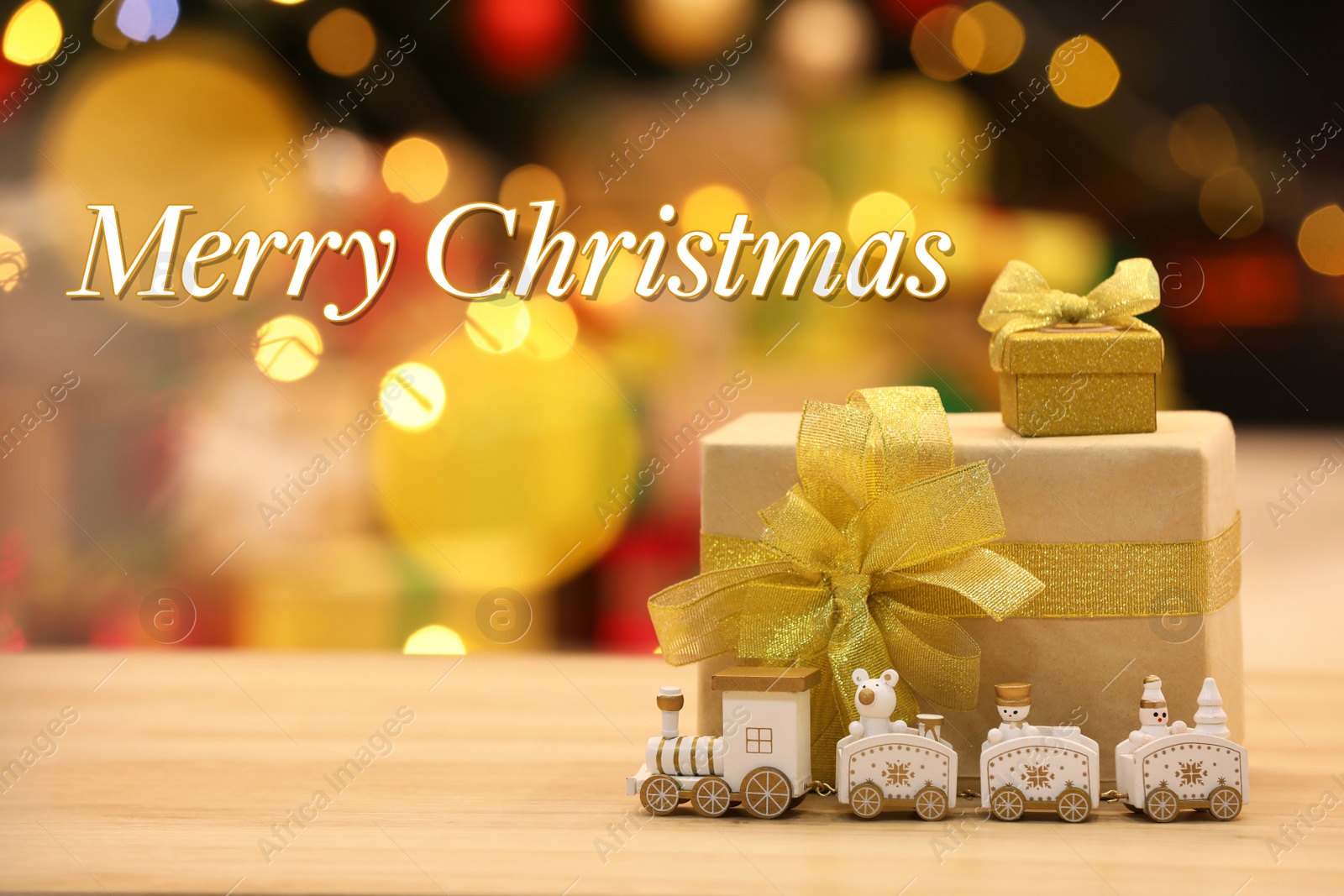 Image of Greeting card with phrase Merry Christmas. Beautiful gift boxes with toy train on wooden table against blurred festive lights