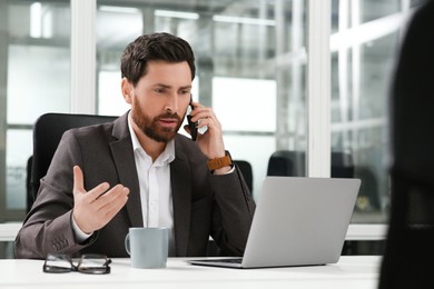 Photo of Man talking on phone while working with laptop at white desk in office