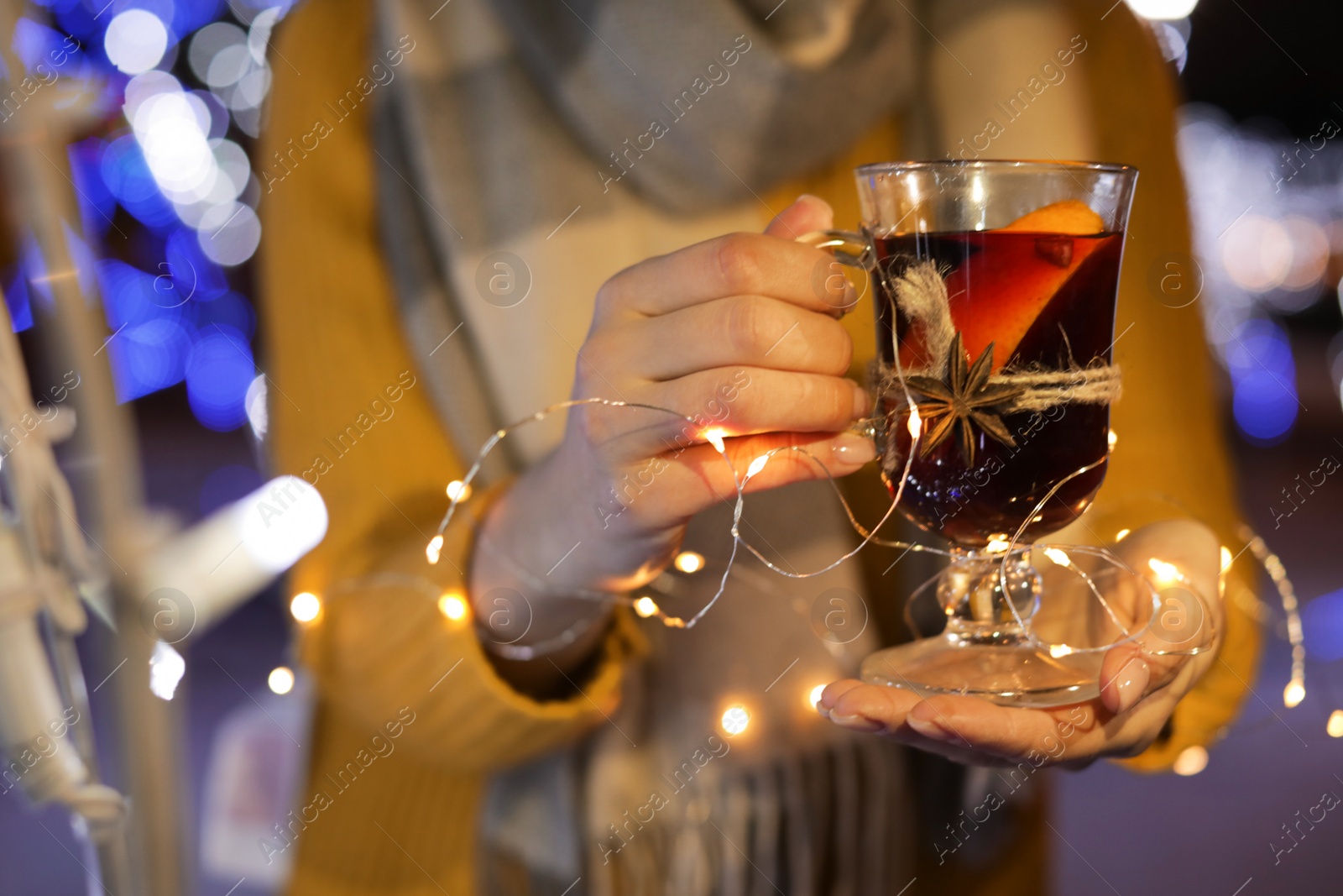 Photo of Woman with glass cup of mulled wine and garland at winter fair, closeup