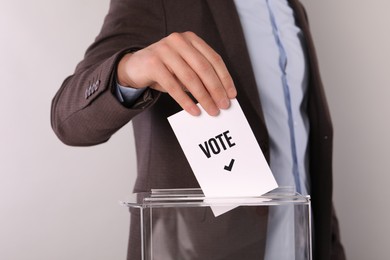 Man putting paper with word Vote and tick into ballot box on light grey background