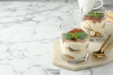 Delicious tiramisu in glasses, mint leaves and spoons on white marble table, space for text