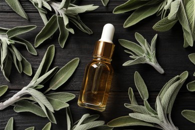 Photo of Bottle of essential sage oil, twigs and leaves on black wooden table, flat lay