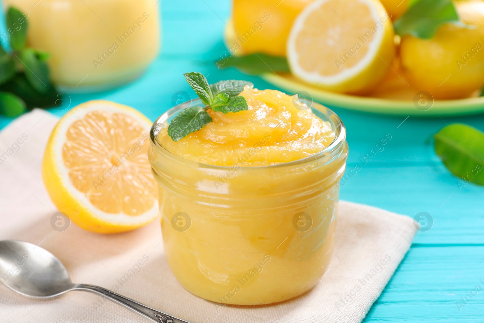 Photo of Delicious lemon curd in glass jar, fresh citrus fruits, mint and spoon on light blue wooden table, closeup