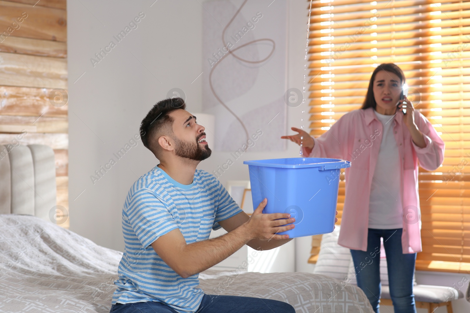 Photo of Young man collecting leaking water from ceiling while his girlfriend calling roof repair service at home