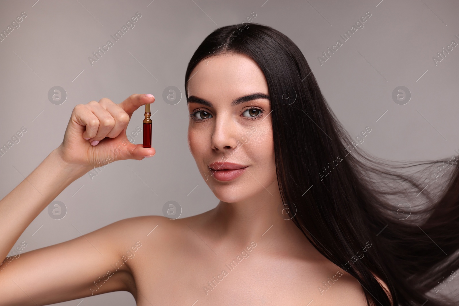 Photo of Beautiful young woman with long healthy hair holding ampoule on grey background