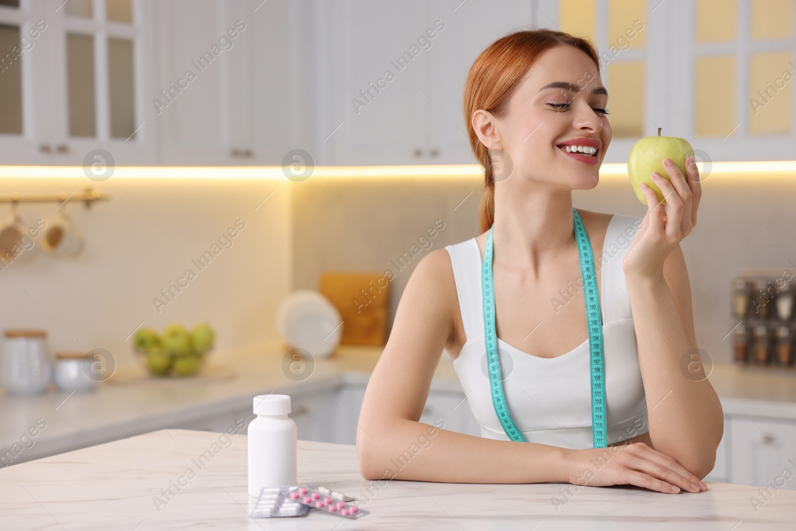 Photo of Young woman with measuring tape and apple at table with pills in kitchen. Weight loss