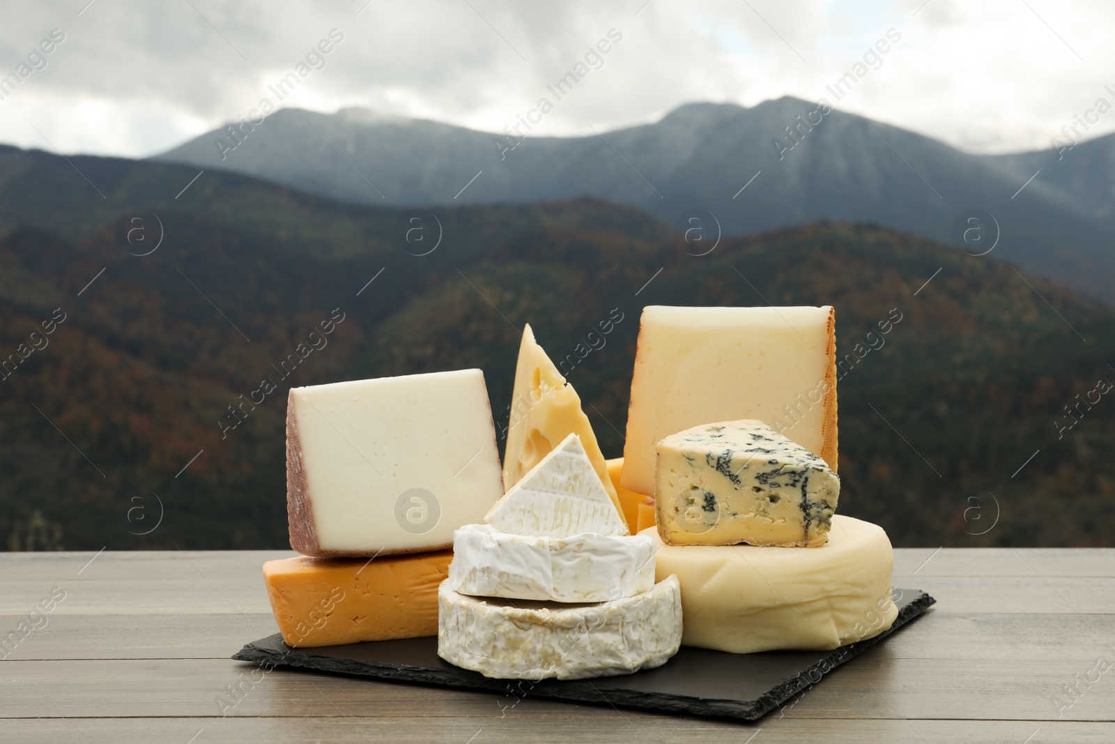 Photo of Different types of delicious cheeses on wooden table against mountain landscape