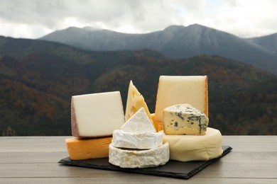 Different types of delicious cheeses on wooden table against mountain landscape