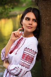 Photo of Beautiful woman in embroidered shirt near tree outdoors. Ukrainian national clothes